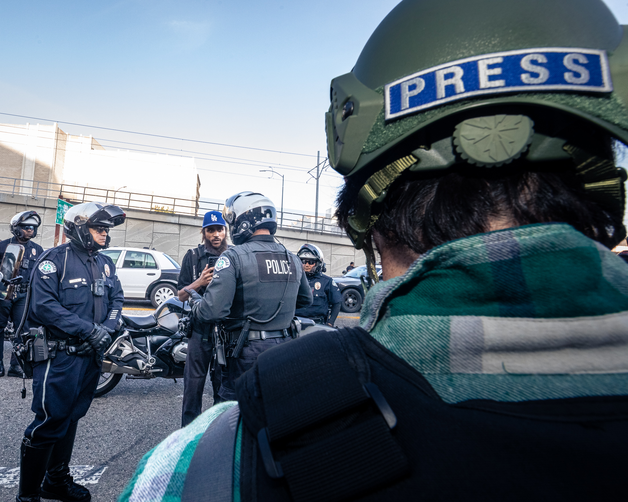 Investigations Newsletter: L.A. TACO Reporter Detained While Reporting (Again) 1 A person wearing a "press" helmet watches two LAPD's speaking to a journalist.